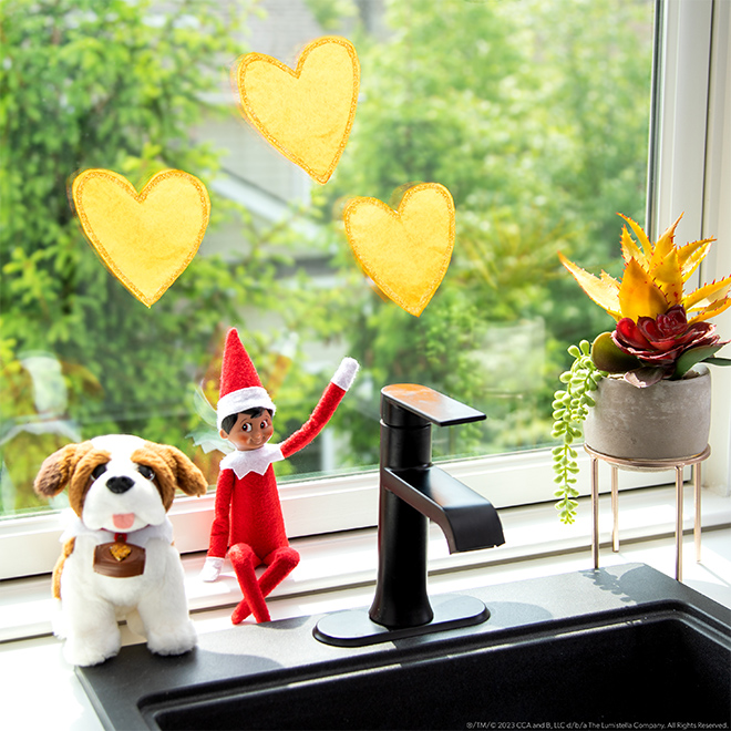 The Elf on the Shelf sits with an Elf Pets St. Bernard in front of a window with DIY suncatcher. 