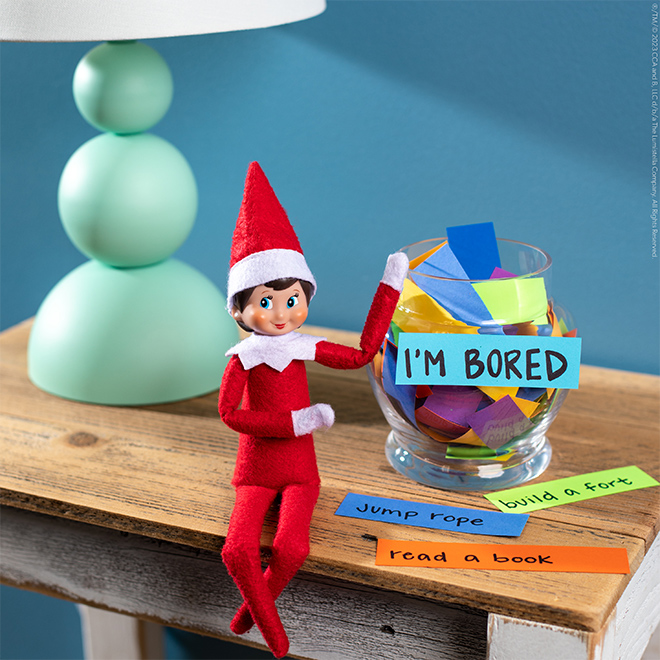 The Elf on the Shelf sitting on a table with a jar filled with activity papers and labeled “I’m Bored”.