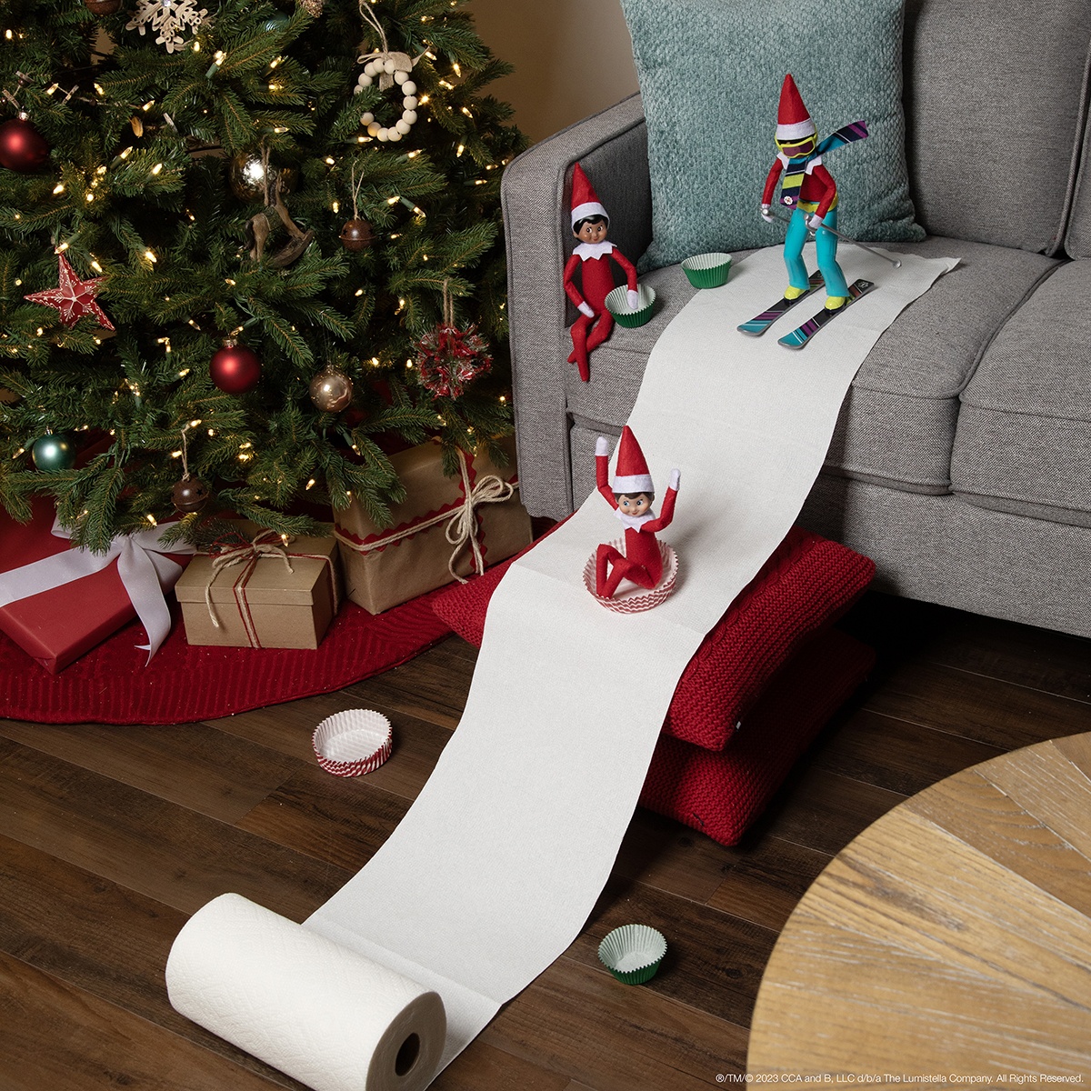 The Elf on the Shelf and friends skiing using a snow hill made of paper towels and sleds made of cupcake wrapping papers