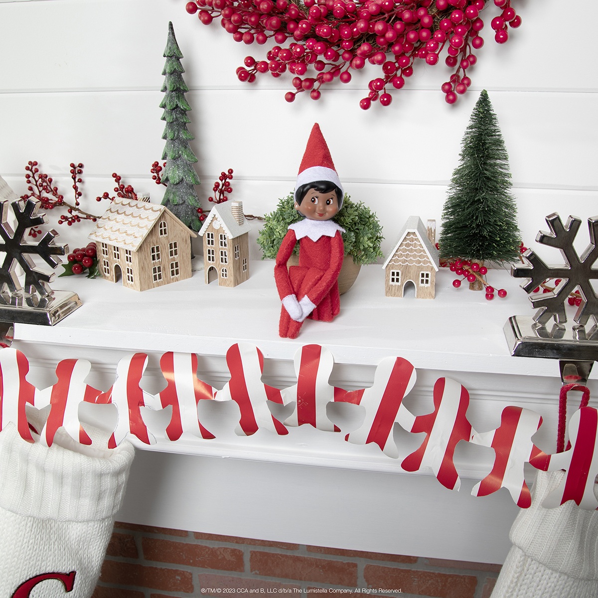 The Elf on the Shelf sitting on a fireplace mantle with garland made from gift-wrapping paper.