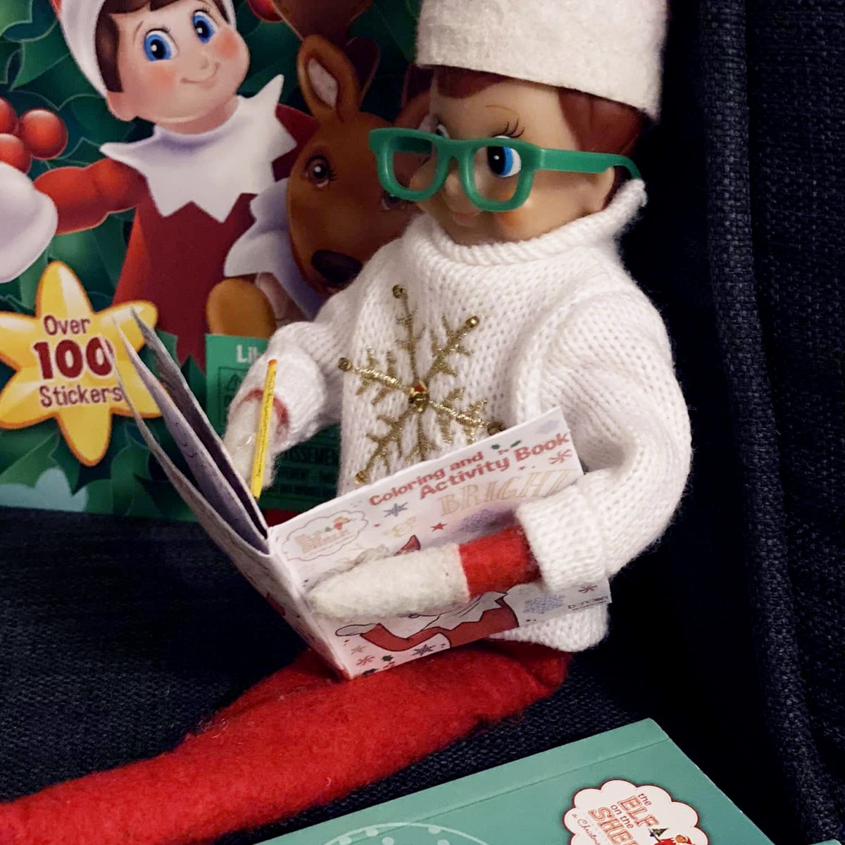 A Scout Elf wearing glasses using a coloring and activity book next to a stack of books