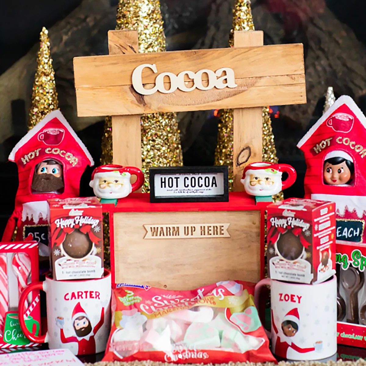 Two Scout Elves in hot cocoa stand costumes next to mugs and hot cocoa bombs with a sign that says "Cocoa, warm up here"