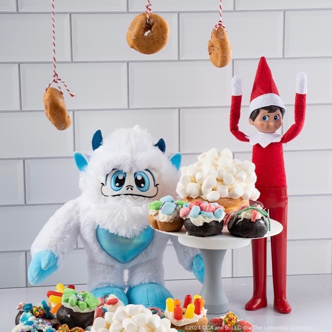 The Elf on the Shelf and a SnoBiggie yeti in front of treats with donuts hanging from string above them