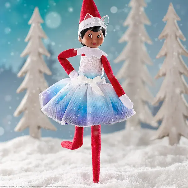 Scout Elf Girl wearing the Noorah dress and ears. She is standing on snow and there is a blue and white winter scene behind her.