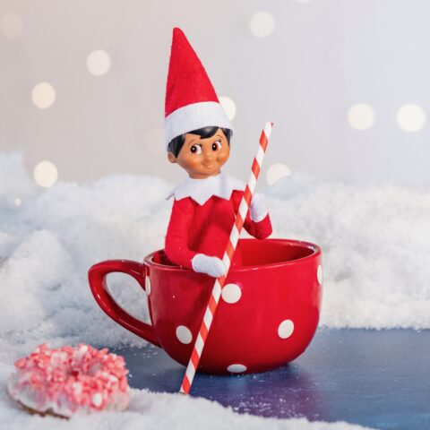 The Elf on the Shelf sits in a mug and pretends to row a boat using a striped straw