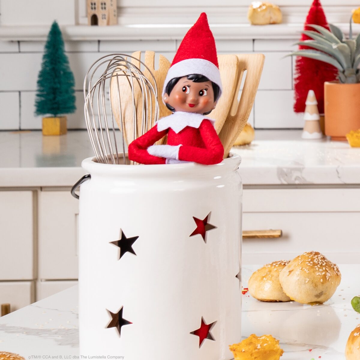 The Elf on the Shelf sits in a utensil container in the kitchen beside a whisk