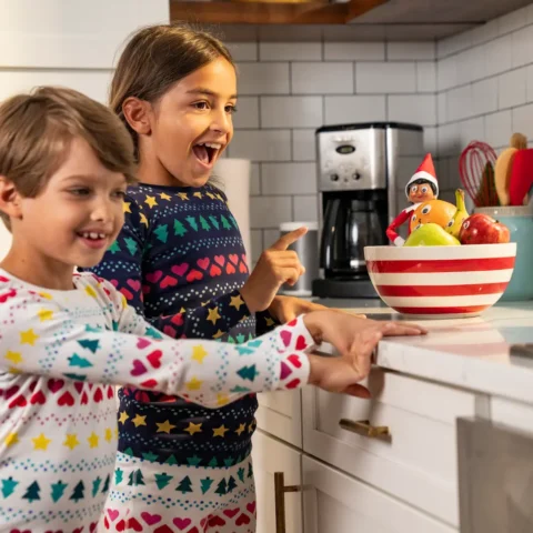 A boy and a girl wearing pajamas discover their Elf on the Shelf sitting in a bowl of fruit