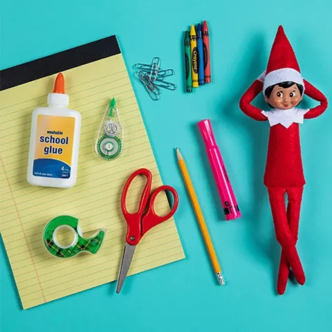 The Elf on the Shelf Scout Elf relaxes next to an assortment of supplies, including paper, crayons, glue, scissors and tape