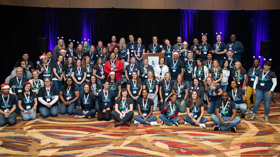 Employees of The Lumistella Company gathered together on a stage around a podium labeled with the Lumiversity logo. Everyone is wearing The Lumistella Company volunteer shirts and have headbands or necklaces with light up stars on them. A few people have Santa hats on. There are black curtains lit by purple lights in the background, and the floor is a red, yellow, orange, and blue geometric pattern.