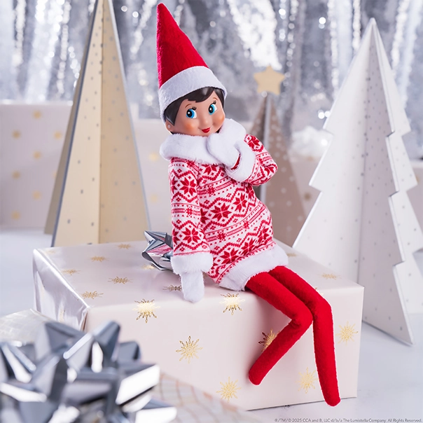 Scout Elf Girl wearing the Claus Couture Collection® Snowflake Sweetie Dress. She is sitting on a wrapped present posing in front of decor conifer trees adorned with festive wrapping paper.