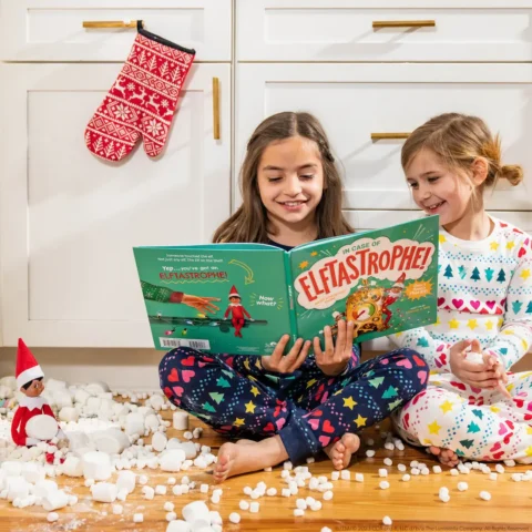 Two girls sit in the kitchen floor reading In Case of Elftastrophe beside The Elf on the Shelf with a mess in the floor