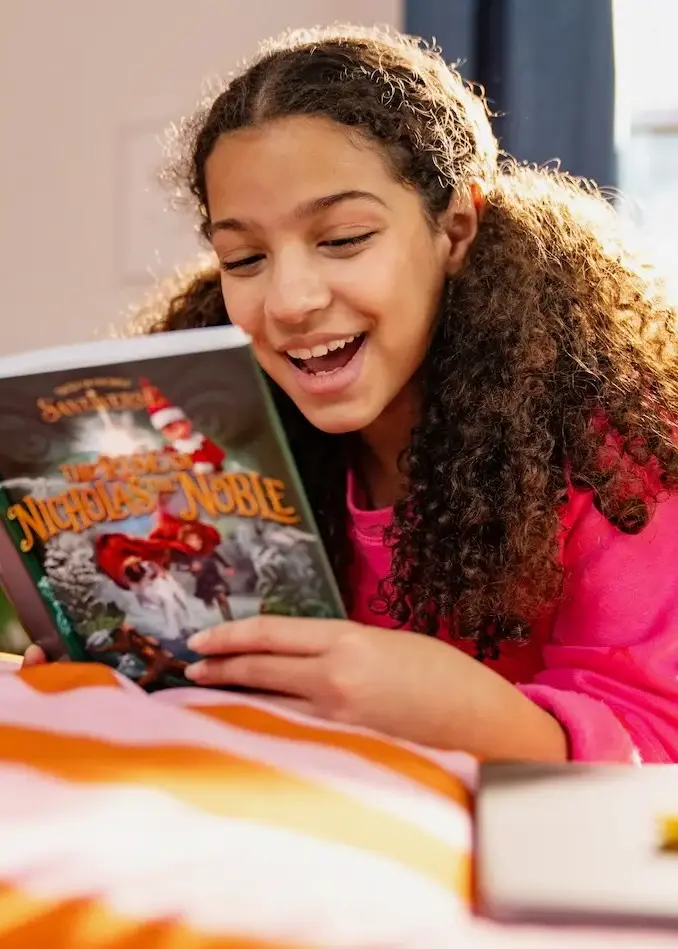 A tween girl reads lays on her stomach on a bed reading The Rise of Nicholas the Noble novel