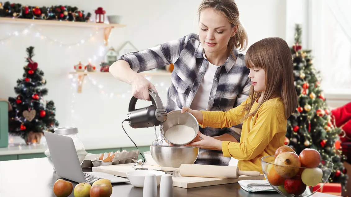 A woman and a young girl baking with each other. They read the recipe from a laptop in front of them on the counter. The woman is holding a hand mixer into a silver bowl while the girl tips a bowl of flower into the mixing bowl. The mixing bowl is on a wooden cutting board and a wooden rolling pin is right next to it. On the counter next to them to the right is a glass bowl filled with pomegranates and apples. On their left is an open box of brown eggs and a clear jar of flour. Next to the laptop is a small white ceramic bowl and salt and pepper shakers. In the background is a white kitchen wall with green cabinets and two wooden floating shelves. There is a smaller Christmas tree on the counter to the left and a larger Christmas tree on the floor on the right. On the higher floating shelf is green Christmas garland. Both shelves contain strings of white lights, and red and green knick knacks.