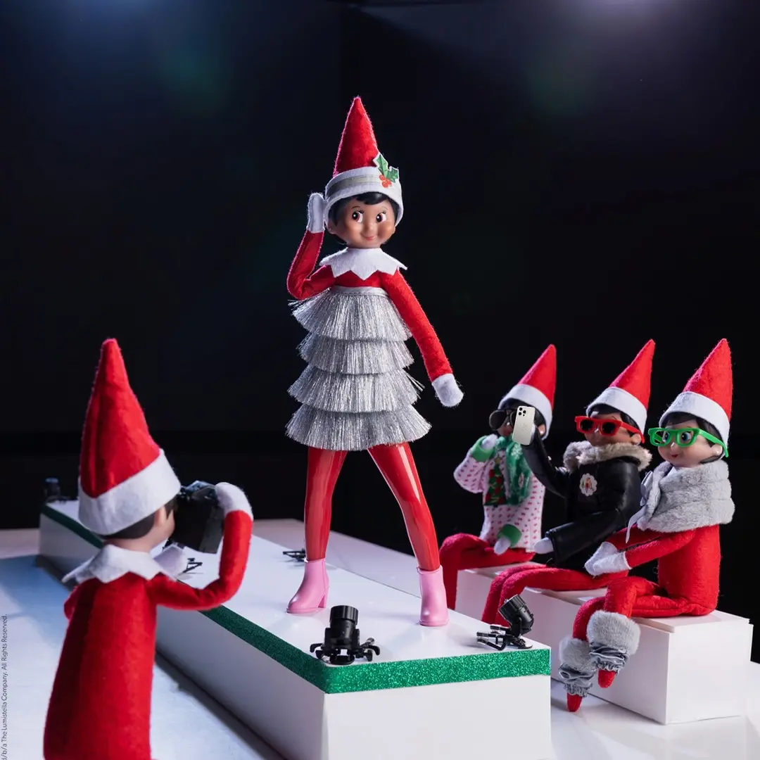 A Scout Elf girl stands on the runway wearing a Tinsel dress as a crowd of elves watches
