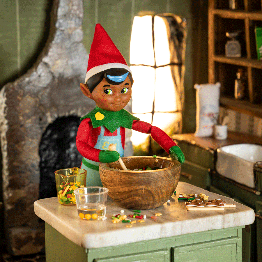 The Elf Mates Chef, in a red hat with a green collar, stands in a kitchen with a fireplace behind him, stirring a bowl of ingredients