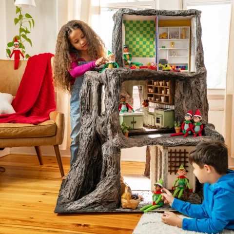 A boy and a girl play with the Elf Mates in a dollhouse shaped like a tree stump