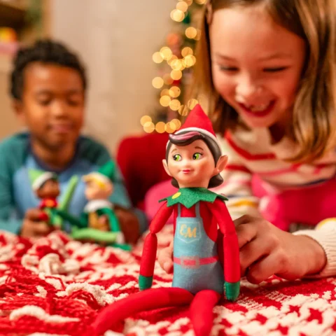 A girl smiles at the Elf Mates Girl Chef, who has a swoosh of pink hair at her forehead, while a boy plays in the background with the Elf Mates Cobbler and Toy Maker
