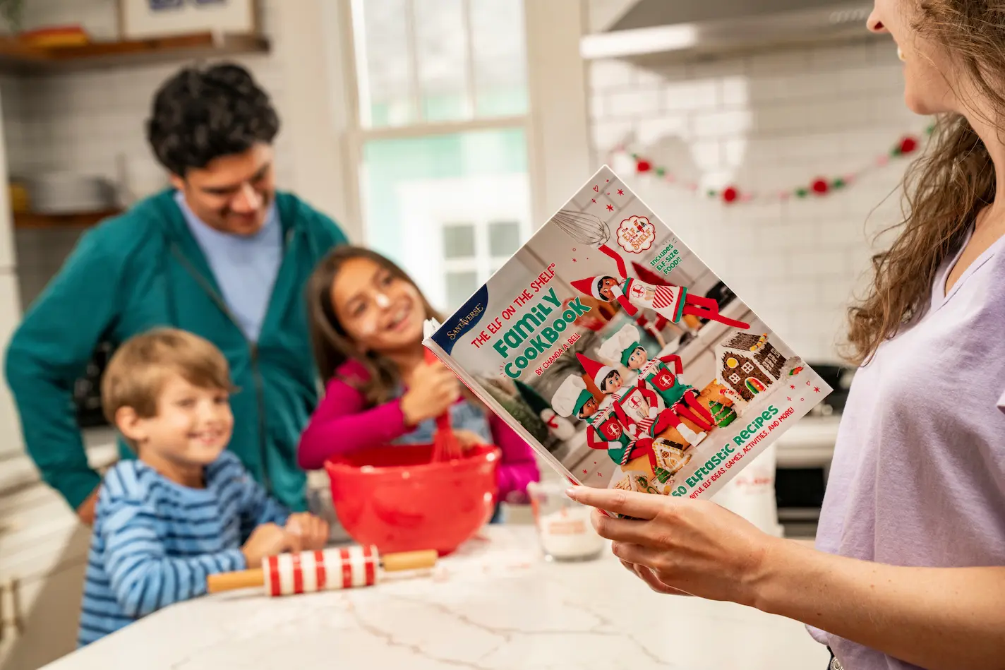 A mom holds The Elf on the Shelf Family Cookbook in the foreground while a dad and two kids mix cookie dough in a bowl