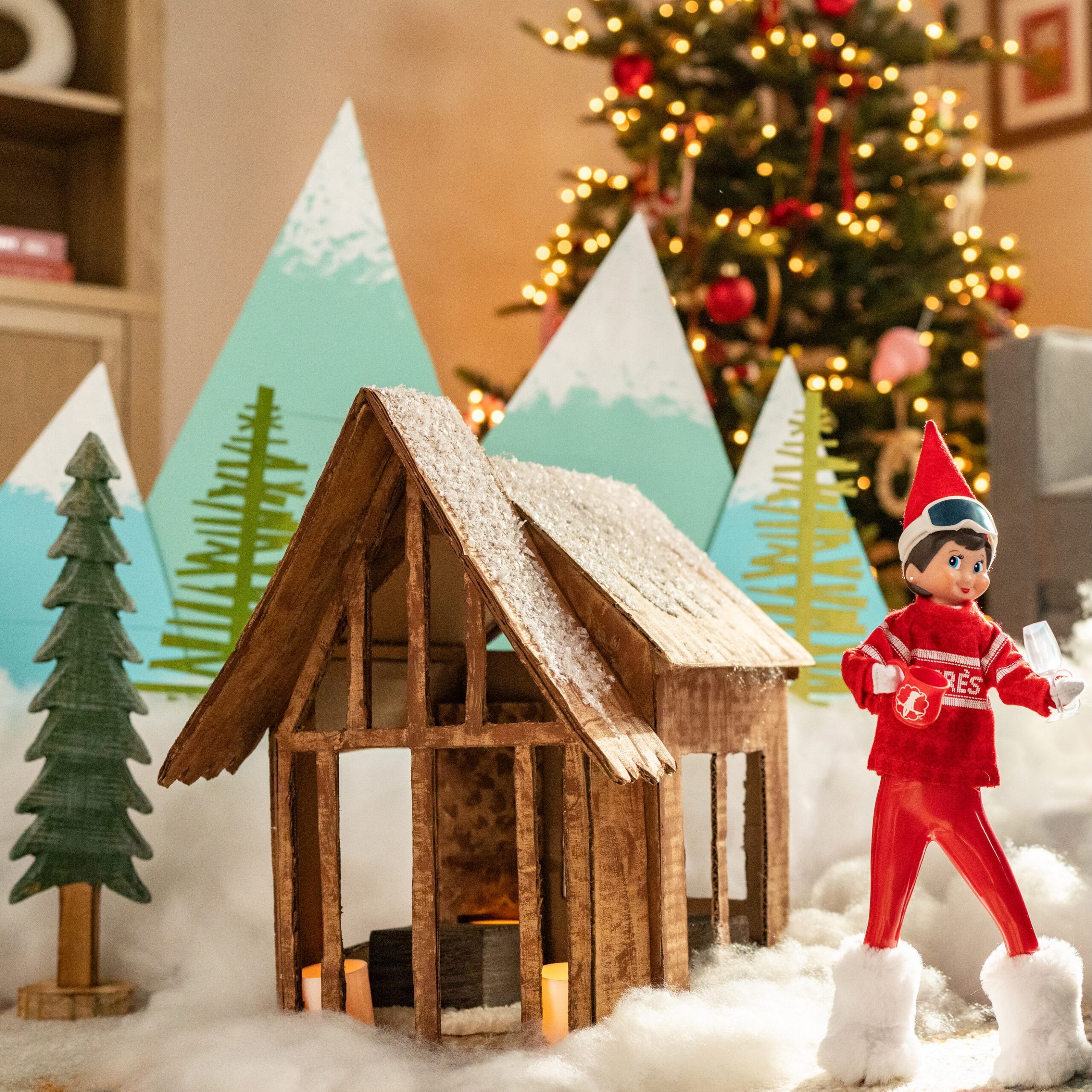 A chic Elf on the Shelf is shown dressed in ski gear in front of a DIY ski chalet with paperboard mountain range