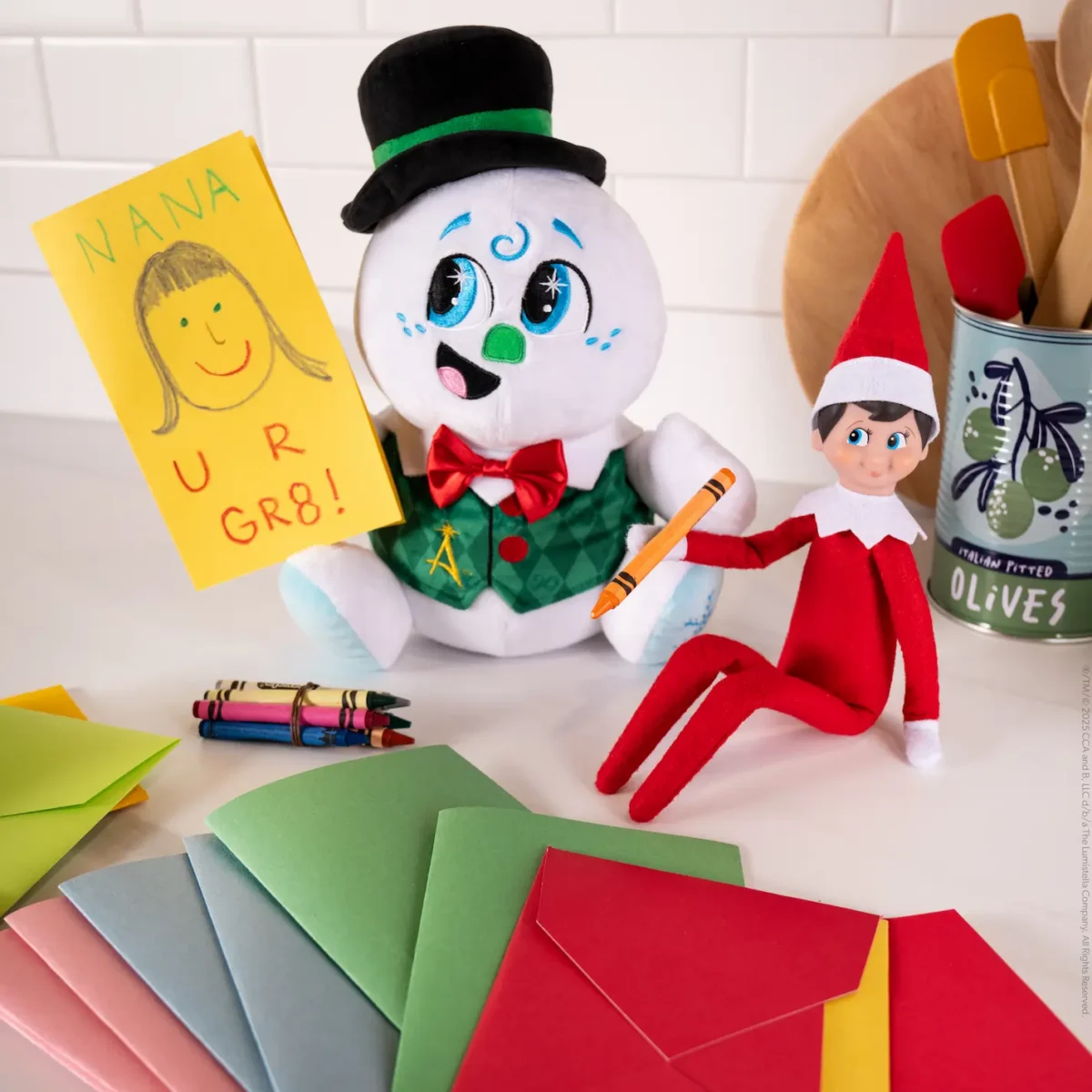 Razzle the Celebration Snowman holds a card with a drawn picture of Nana and written with the words "U R Gr8" while the Scout Elf holds a crayon