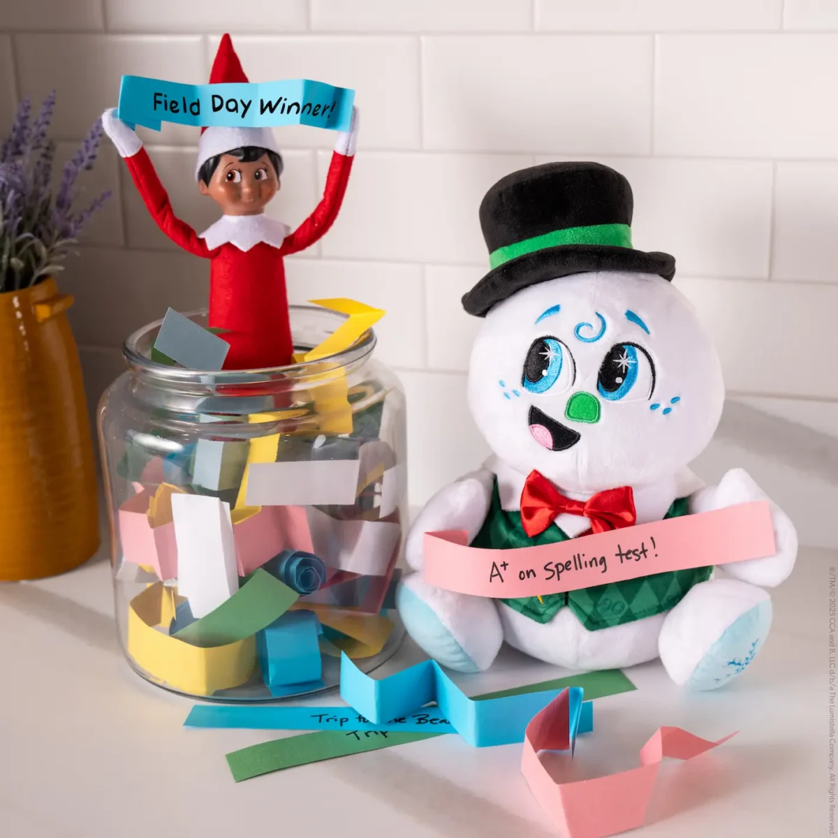 Razzle the Celebration Snowman holds a slip of pink paper that says "A+ on Spelling Test" and The Elf on the Shelf Scout Elf holds a slip of blue paper that says "Field Day Winner"