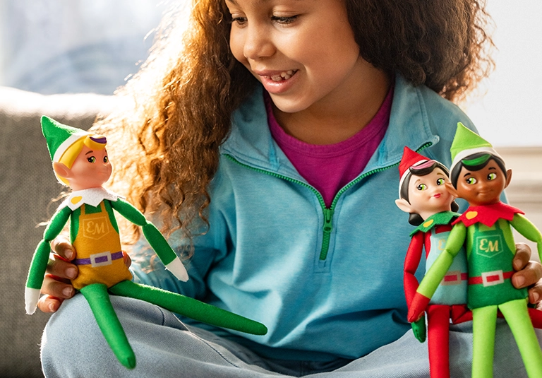 A young girl is playing with the Elf Mates® Toy Maker, Elf Mates® Chef and Elf Mates® Cobbler. She appears to be seated on a sofa in her living room. She is backlit by the bright, natural light of the sun, shining into the room, accentuating her curly brown hair.