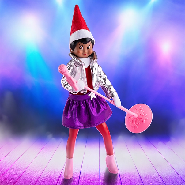 A Scout Elf is gripping the mic for her next big performance! She is adorned in the pants, jacket and skirt included in the MagiFreez® Pop Star set, of which the microphone is also included. The background is a brightly lit stage with cyan and magenta lighting.
