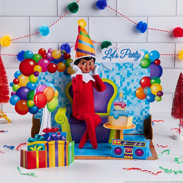 A Scout Elf wearing a party hat is posed in one of the included scenes from the Insta-Moment™ Pop Ups, Celebration Series. The pop up says "Let's Party" and there are balloons and wrapped presents with a boom box. The scene appears to be a birthday party setting and there are pom-poms strewn about in the background.
