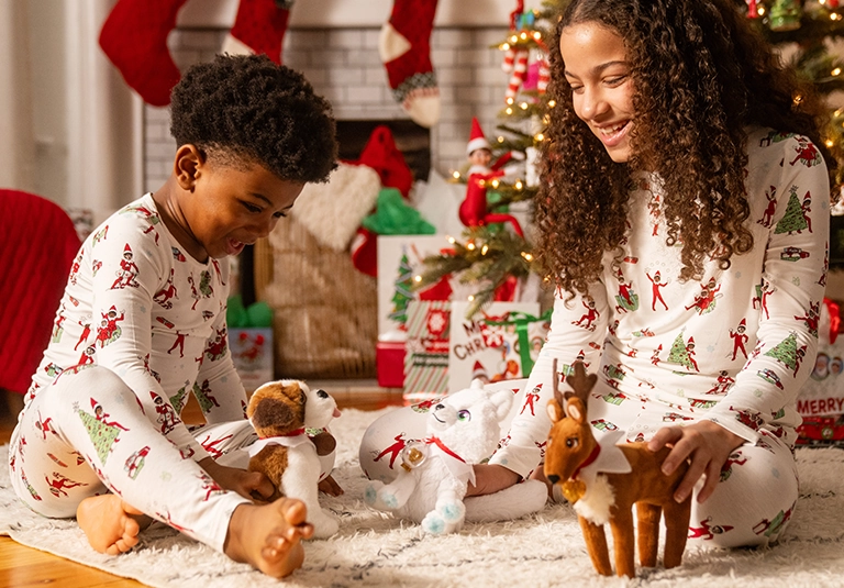 A pair of siblings are playing with the Elf Pets® Saint Bernard, Arctic Fox and Reindeer. The brother has the Saint Bernard and the sister has the Arctic Fox and Reindeer. They are seated on the floor, playing on a white rug. They are both dressed in festive, Christmas pajamas. Behind them is a festively decorated living room in preparation for Christmas. There are presents and a Christmas tree, with stockings hanging from the fireplace.