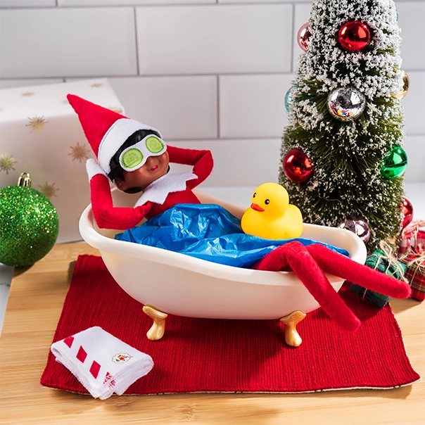 A Scout Elf relaxes in the elf-size bathtub from the Polar Props™ S’elf Care Set, wearing the included cucumber mask. Blue craft paper is made to look like water in the bathtub. The included rubber ducky floats, while the with the tiny bath towel folded on the ground. There is a conifer decor tree with ornaments in the background.