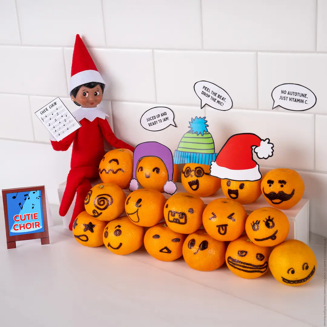A Scout Elf sits beside a stack of tangerines with faces drawn on them, arranged as a choir as if they are singing.