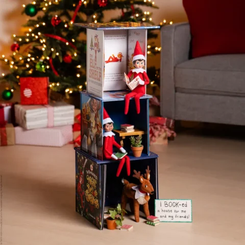 Two Elf on the Shelf elves and an Elf Pets Reindeer stand near a three-level house made of books