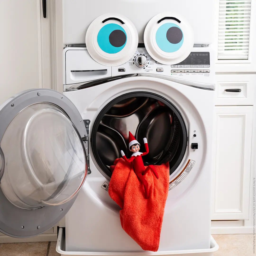 A Scout Elf sitting on a frontloading washing machine with its door open, red cloth spilling out of the opening as if it is a tongue and two googly eyes placed at the top
