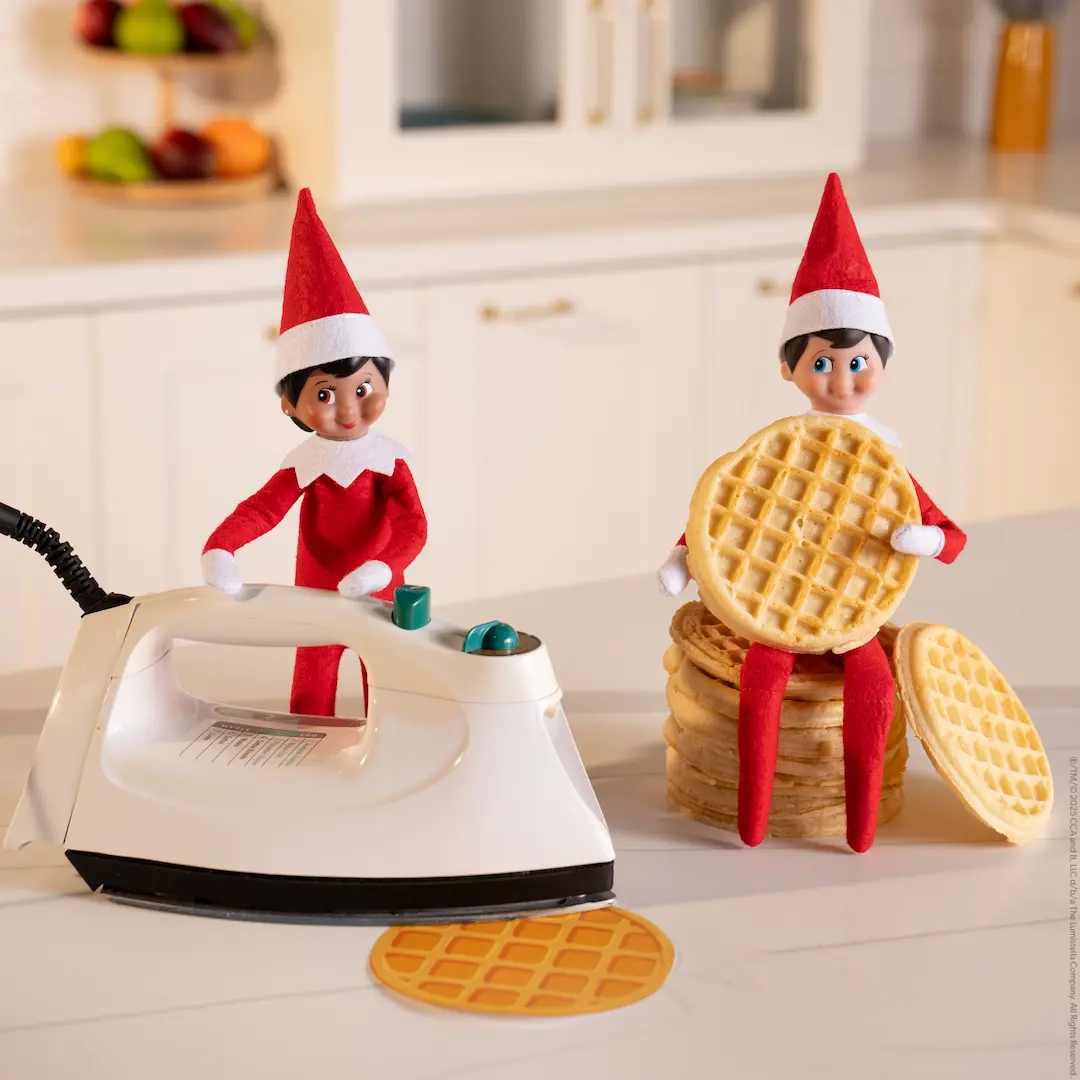 A Scout Elf pretends to iron a waffle flat while another elf holds a waffle