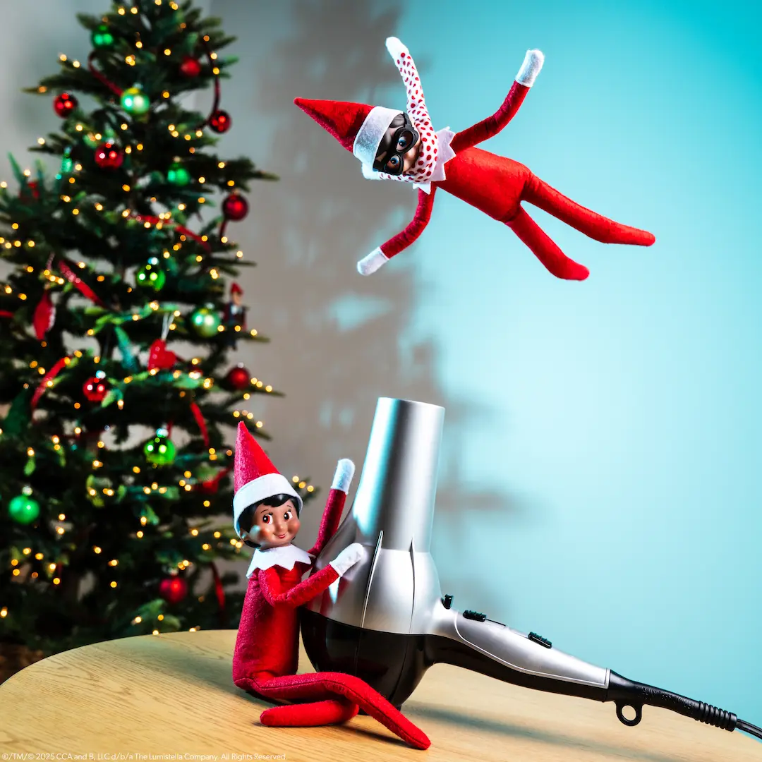 The Elf on the Shelf Scout Elf girl holds a hair dryer while The Elf on the Shelf Scout Elf boy practices flying in the stream of air