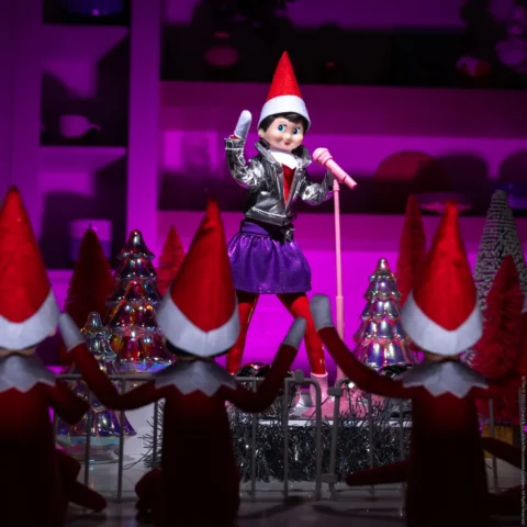 The Elf on the Shelf Scout Elf girl stands on a mini stage in front of a microphone as if performing at a concert