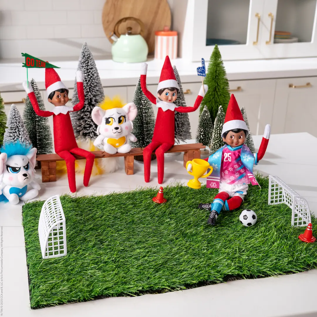 The Elf on the Shelf kicks a ball on a mini soccer field while other Scout Elves and Bogie characters watch