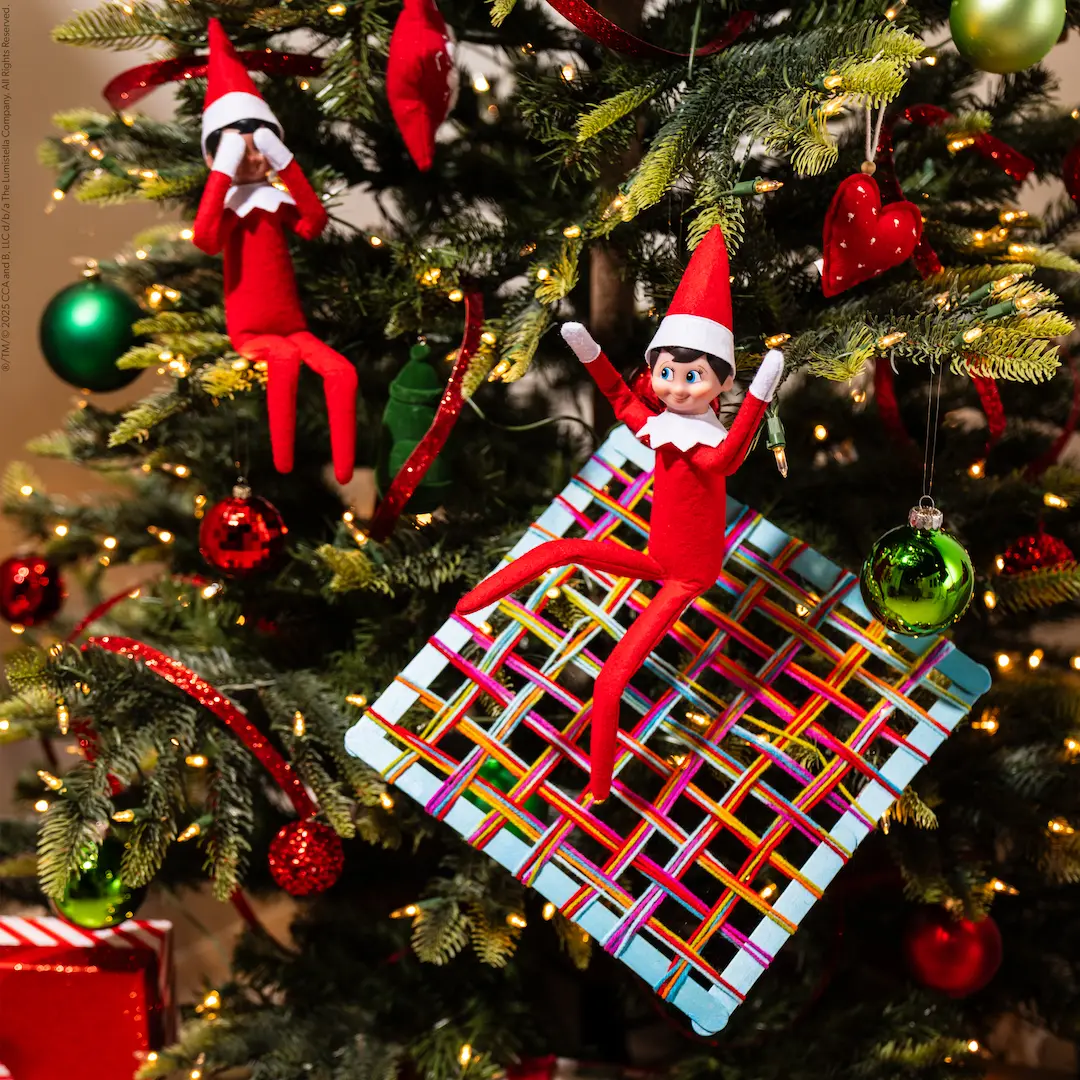 The Elf on the Shelf lands on a handmade net stuck in the Christmas tree as another elf watches