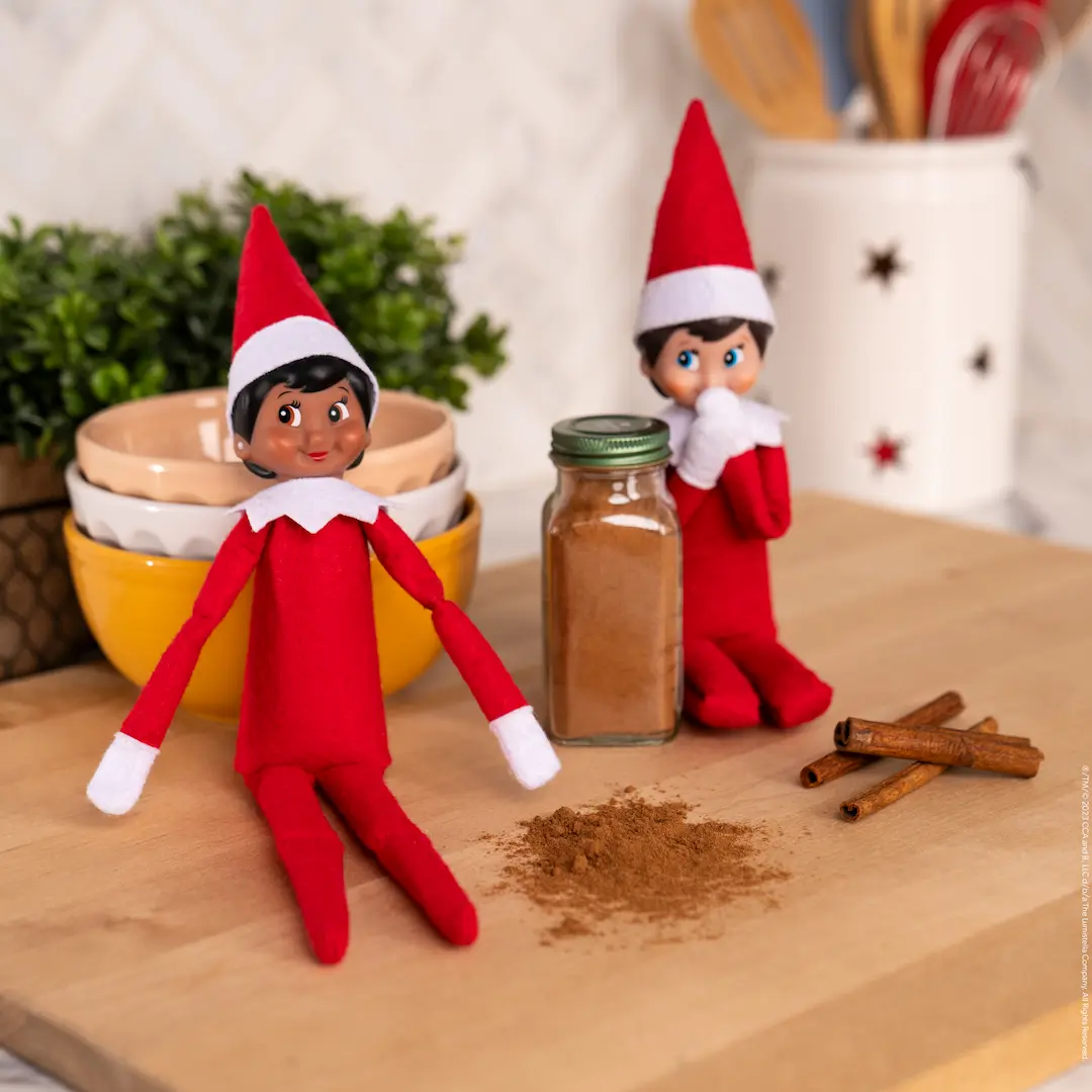 Two Scout Elves sit beside a jar of cinnamon with a little sprinkled out beside them