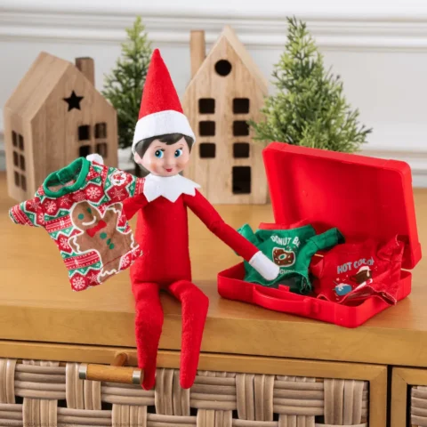 The Elf on the Shelf Scout Elf girl sits beside a packed suitcase of elf clothing