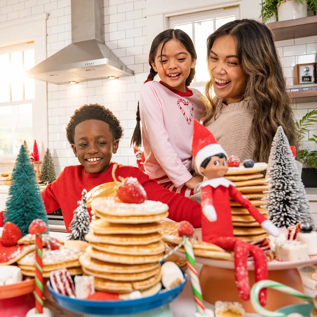A mom and two children stand behind a table set with stacks of pancakes for a North Pole breakfast as The Elf on the Shelf sits nearby watching