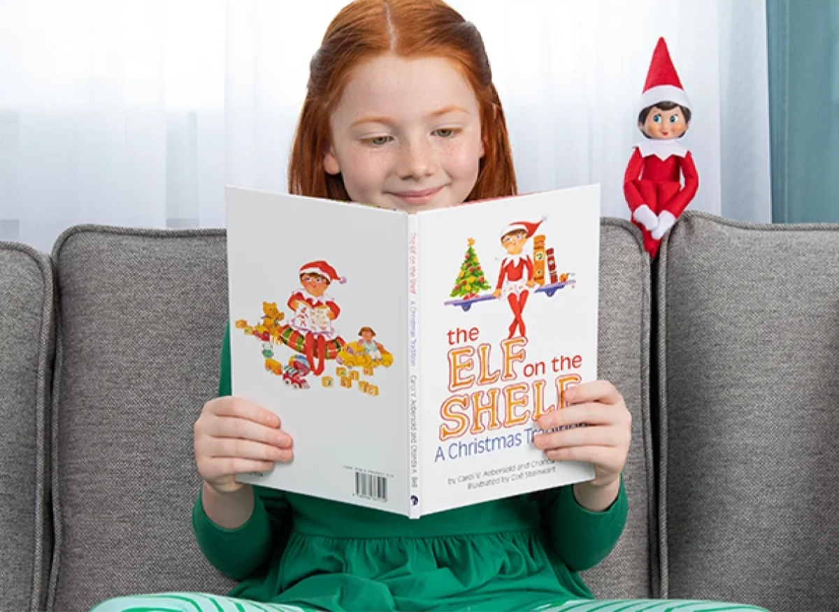 A young girl reads The Elf on the Shelf®: A Christmas Tradition, seated on a sofa. To her left, her Scout Elf also sits on the top of the sofa, overlooking her.