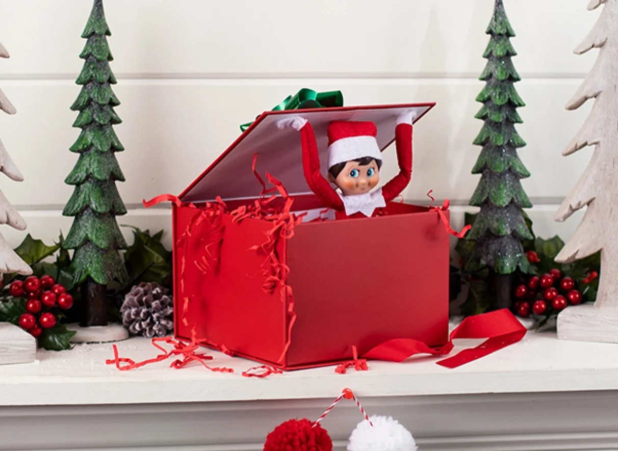 A Scout Elf is posed inside a festive, red gift box with crinkle-cut paper shreads bursting out of the box. The Scout Elf is inside the box, lifting the lid with his arms. The gift box appears to be on the mantle of a fireplace. Also pictured alongside the gift box are a pair of wooden, decorative conifer trees.