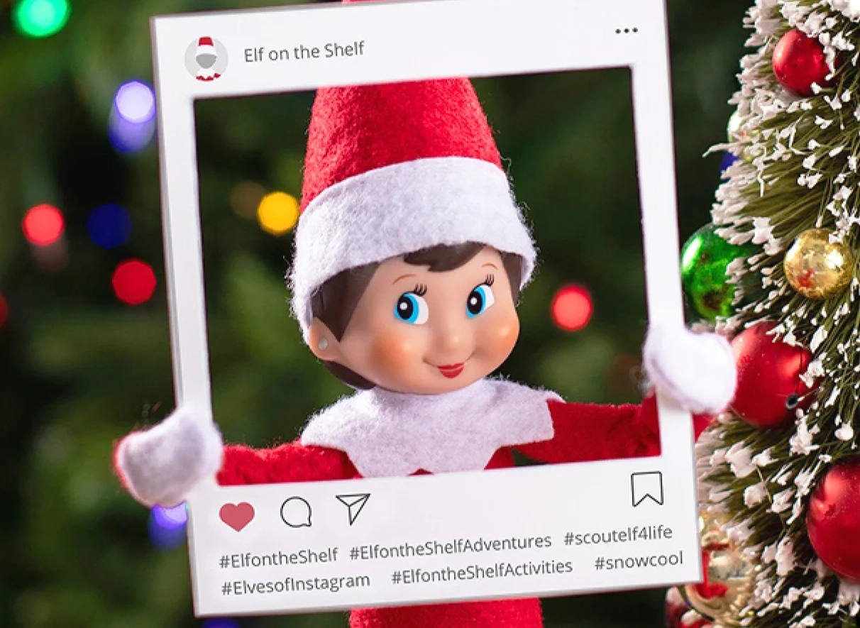 A Scout Elf is posed holding with an Instagram-inspired frame that makes it look like she is a social media post. In the background, just out of focus is a decorated Christmas tree with colored Christmas lights.