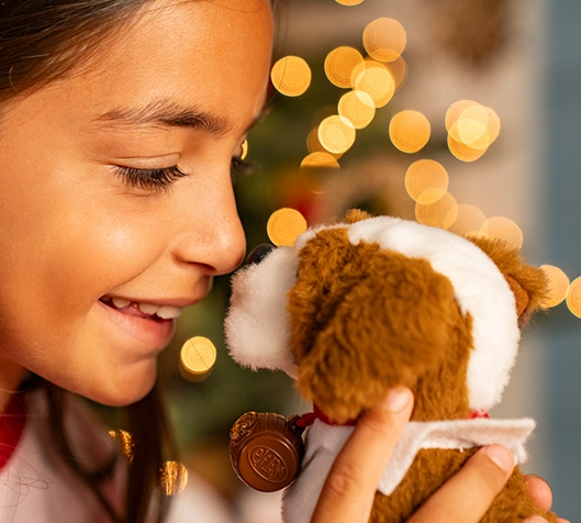 A young girl clutches her Elf Pets® St. Bernard pup plush and nuzzles him with her nose. In the background, you can make out a festive, decorated and lit Christmas tree.