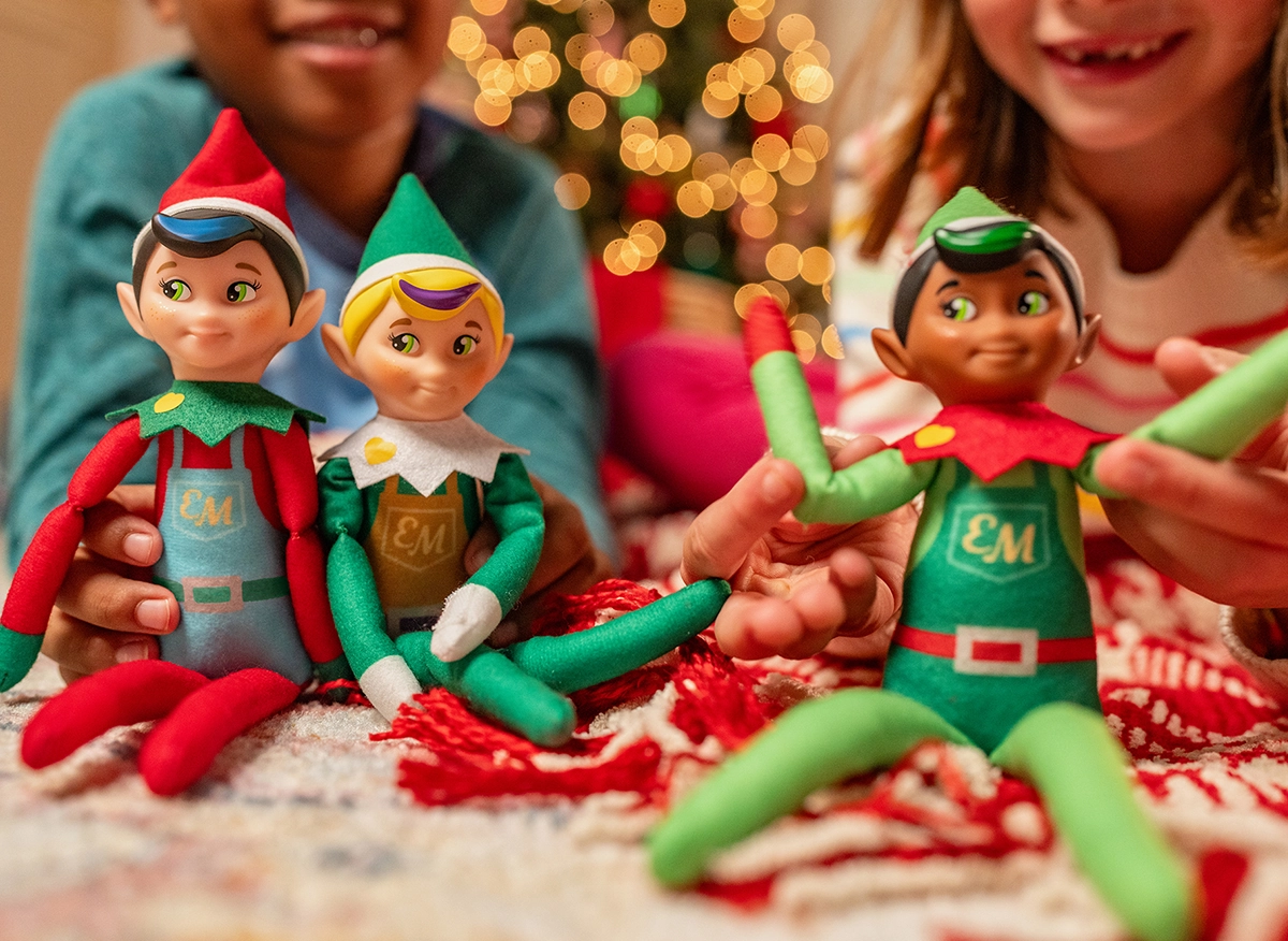 A pair of children play with a trio of Elf Mates: the Chef, Toy Maker and Cobbler. In the background you can make out a blurry Christmas tree. The children are situated on the floor playing with the Elf Mates dolls.
