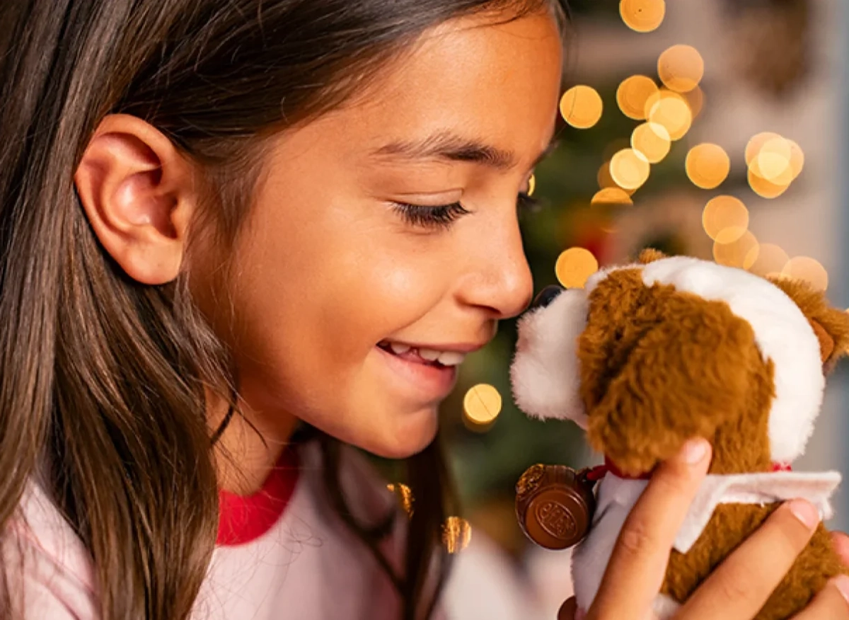 A young girl nuzzles her Elf Pets® Saint Bernard pup. Christmas lights are blurred in the background.