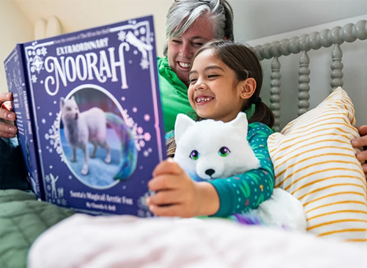 A grandmother and her granddaughter are lying in bed reading the "Extraordinary Noorah" book. The granddaughter is cuddling with the Noorah plush.