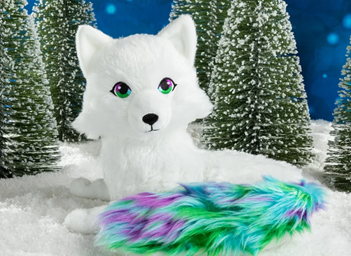 The Noorah plush is posing in a winter wonderland setting with powdery snow, snow-capped conifer trees and a dark blue background showing a light snowfall.