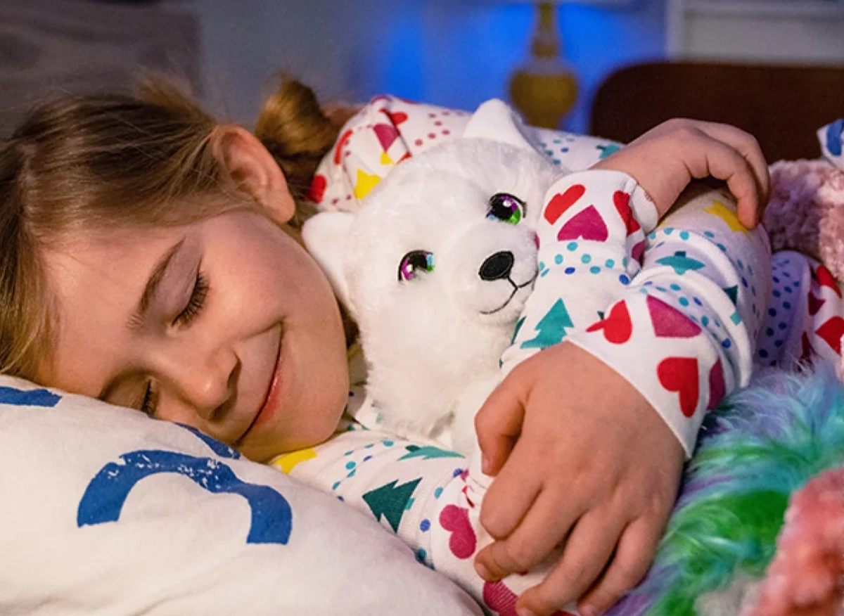 A young girl is pictured snuggling her Noorah plush in bed. Her eyes are closed and she is smiling.